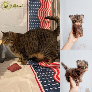May include: A brown tabby cat with green eyes stands on a white surface with a red, white, and blue American flag. The cat is looking to the left. A small brown and black amigurumi cat is being held in a hand. The amigurumi cat has green eyes and a fluffy texture.