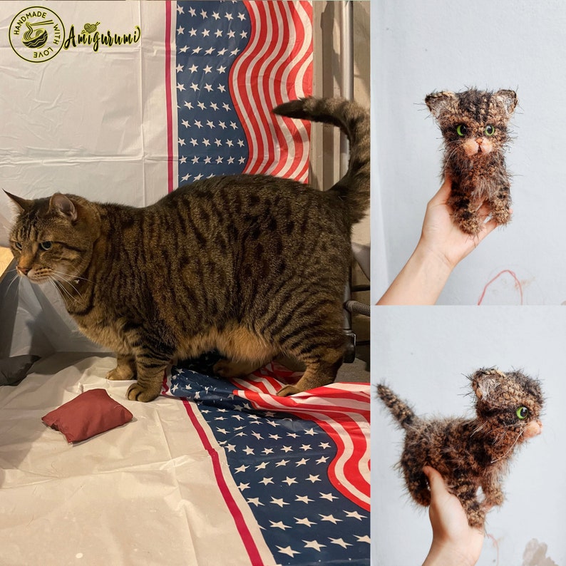 May include: A brown tabby cat with green eyes stands on a white surface with a red, white, and blue American flag. The cat is looking to the left. A small brown and black amigurumi cat is being held in a hand. The amigurumi cat has green eyes and a fluffy texture.