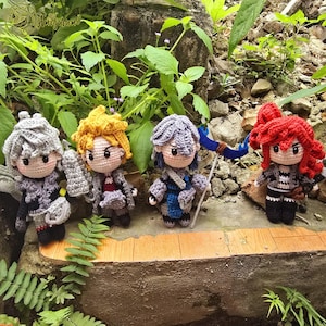 May include: Four handmade crochet dolls with detailed features. Each doll has a unique hairstyle and outfit, including armour and weapons. The dolls are displayed outdoors among greenery, showcasing the craftsmanship and character design.