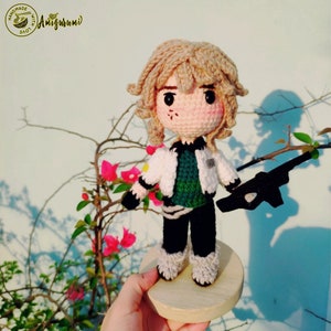 May include: Crocheted amigurumi doll with blonde hair, wearing a white jacket, green shirt, black pants, and holding a black gun. The doll is standing on a wooden base.
