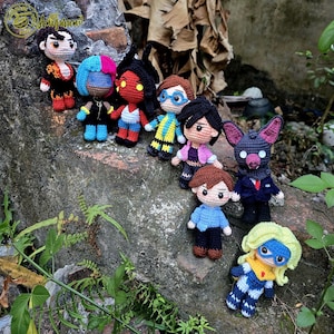 Puede incluir: Una colección de muñecos de ganchillo hechos a mano, cada uno con diseños de personajes únicos. Los muñecos presentan varios atuendos y accesorios, mostrando una gama de colores y estilos. Los muñecos están dispuestos sobre una superficie natural texturizada.