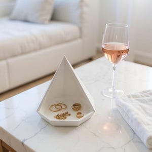 Peut inclure: Un plateau à bijoux géométrique blanc contient des bagues et des colliers en or. Un verre de vin rosé et une serviette blanche sont posés sur une table en marbre. Le plateau est en forme de pyramide.