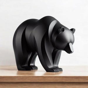 May include: A matte black bear sculpture with a modern, geometric design. The bear is standing on all fours, with its head slightly lowered. The sculpture is placed on a wooden surface.