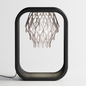 May include: A modern table lamp with a black rectangular frame. The lamp features a white, intricate, organic-shaped shade. The shade's design allows light to filter through, creating a unique lighting effect. The lamp is plugged into a wall outlet.