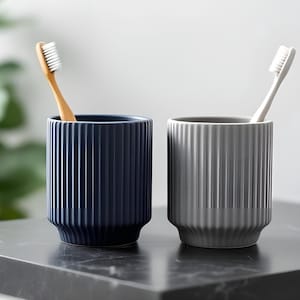 May include: Two ceramic toothbrush holders, one navy blue and one gray, with vertical ribbed detailing. Each holder contains a toothbrush. The navy blue holder has a wooden toothbrush, and the gray holder has a white toothbrush.