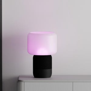 May include: A black smart speaker with a white frosted glass lampshade that emits a pink light. The speaker has a black mesh grille.