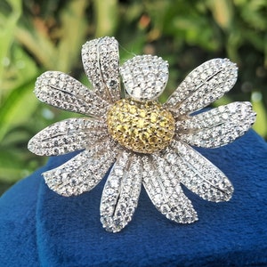 Puede incluir: Un broche de plata en forma de margarita con un centro amarillo, engastado con piedras blancas brillantes. El broche se muestra sobre un fondo de terciopelo azul.