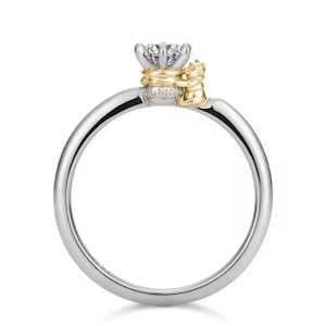 May include: A silver ring featuring a gold crown and a small figure holding the crown. The ring is set with a single diamond.