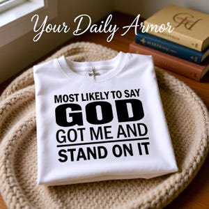 May include: A folded white t-shirt with the text "MOST LIKELY TO SAY GOD GOT ME AND STAND ON IT" in black. The shirt is on a beige knitted surface. The words "Your Daily Armor" are written in cursive at the top.