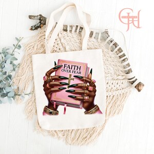 May include: White tote bag featuring a graphic of hands adorned with rings holding a pink book with the words "FAITH OVER FEAR" and a gold cross. The hands have long black fingernails. The bag has cream-colored handles.