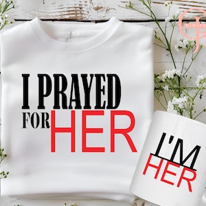 Puede incluir: Camiseta blanca con texto negro que dice "Rezé por" y texto rojo que dice "ELLA". Una taza blanca con texto negro que dice "Soy" y texto rojo que dice "SUYA".