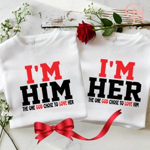 May include: Two white t-shirts with the phrases "I'M HIM" and "I'M HER" in red and black lettering. The text "THE ONE GOD CHOSE TO LOVE HER" and "THE ONE GOD CHOSE TO LOVE HIM" are below the main text. A red bow and rose are also in the image.