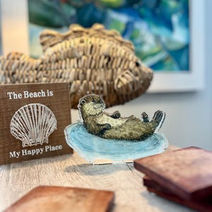 May include: A wooden sign with the text "The Beach is My Happy Place" next to a decorative otter figurine lying on a blue surface. A woven fish-shaped decoration and a painting are in the background.