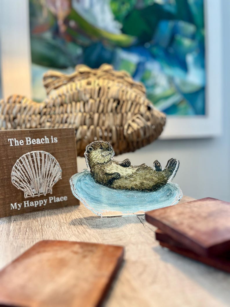 May include: A wooden sign with the text "The Beach is My Happy Place" next to a decorative otter figurine lying on a blue surface. A woven fish-shaped decoration and a painting are in the background.