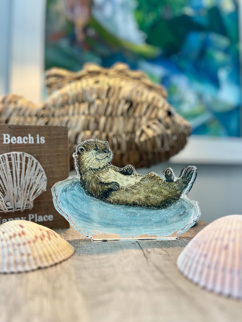 May include: A decorative otter figurine resting on a blue wave-shaped base. The otter is brown and tan, with a relaxed pose. Seashells and a wooden sign that says "Beach is" are also visible, suggesting a coastal theme.