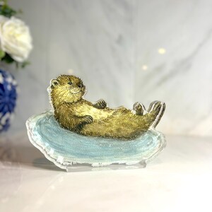 May include: A clear acrylic figurine of a relaxed otter lying on a blue, wave-shaped base. The otter has brown fur and is positioned with its paws up. The figurine is displayed on a white surface.