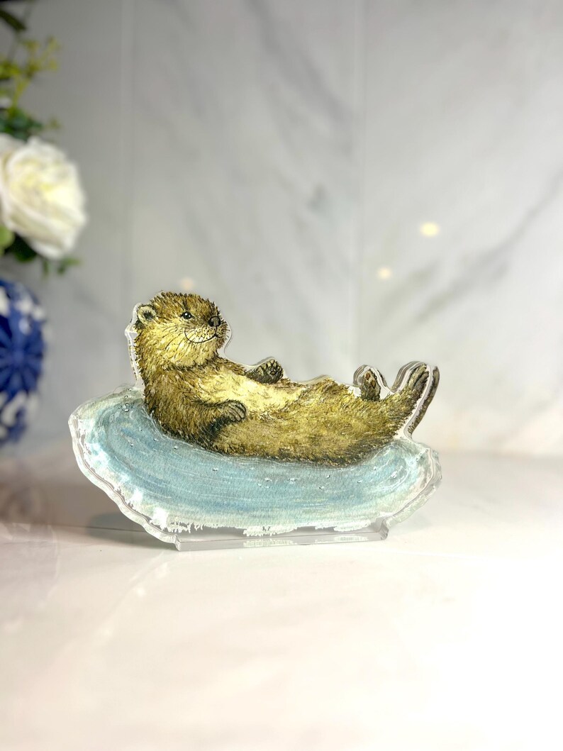 May include: A clear acrylic figurine of a relaxed otter lying on a blue, wave-shaped base. The otter has brown fur and is positioned with its paws up. The figurine is displayed on a white surface.