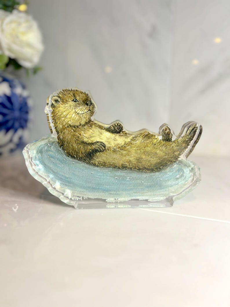 May include: A clear acrylic figurine of a relaxed otter lying on a blue, cloud-shaped base. The otter is depicted with brown fur and a smiling expression. The figurine is displayed on a clear stand, suitable for a desk or shelf.