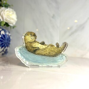 May include: A clear acrylic figurine of a relaxed otter lying on a light blue surface. The otter has brown fur and is positioned horizontally. The figurine is a decorative item.