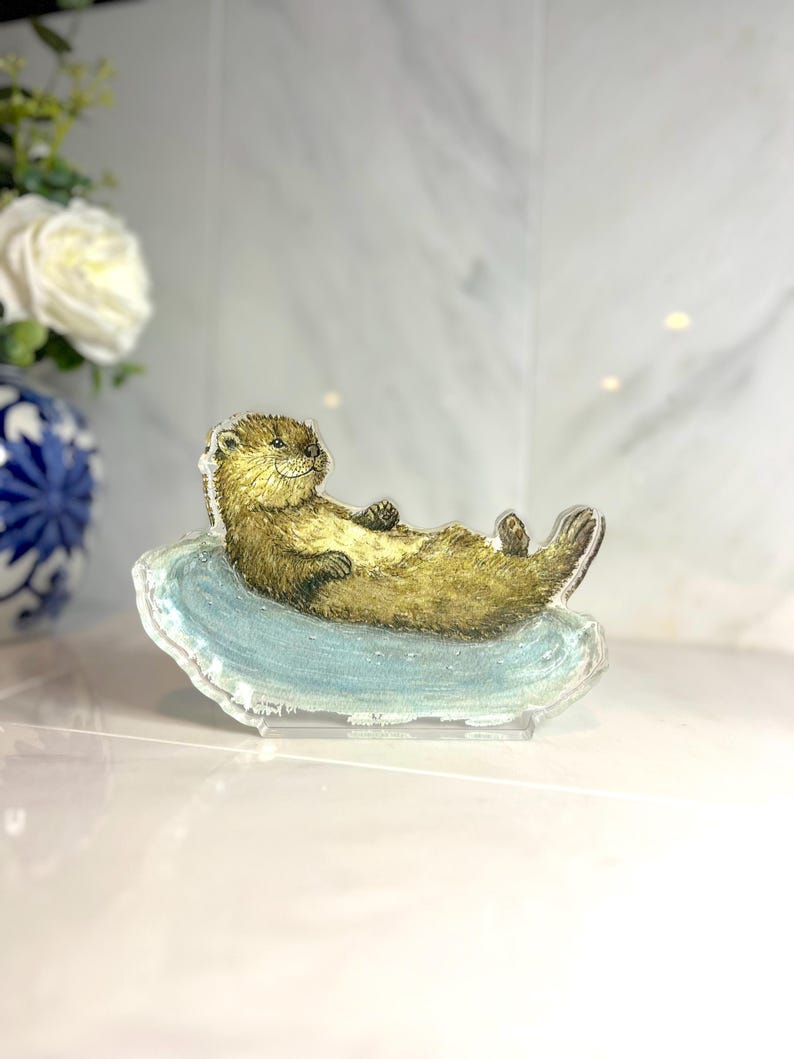 May include: A clear acrylic figurine of a relaxed otter lying on a light blue surface. The otter has brown fur and is positioned horizontally. The figurine is a decorative item.
