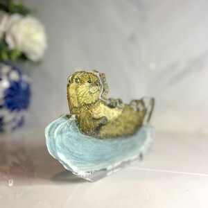 May include: A detailed figurine of an otter resting on a light blue, wave-shaped base. The otter has brown fur and is lying on its back. The base is translucent, and the background is blurred.