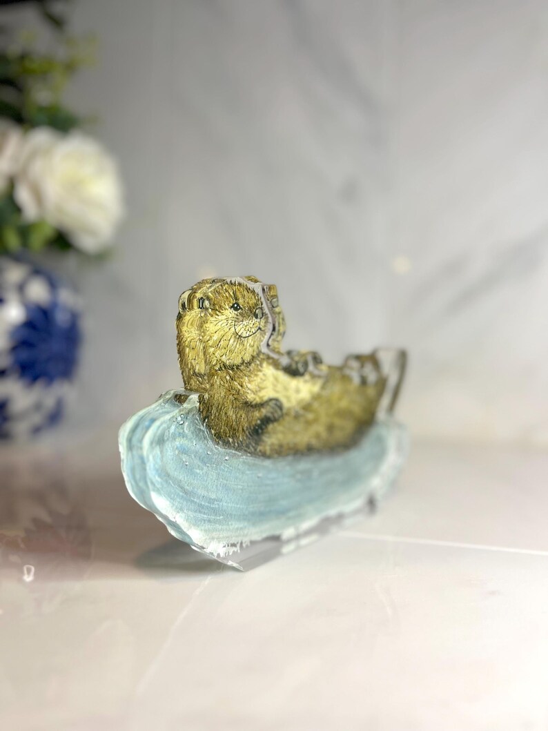 May include: A detailed figurine of an otter resting on a light blue, wave-shaped base. The otter has brown fur and is lying on its back. The base is translucent, and the background is blurred.