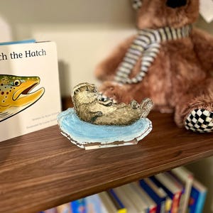 May include: A detailed figurine of a sea otter resting on a light blue base. The otter is brown with a light belly, and the base has a white border. A children's book and a brown teddy bear are in the background.