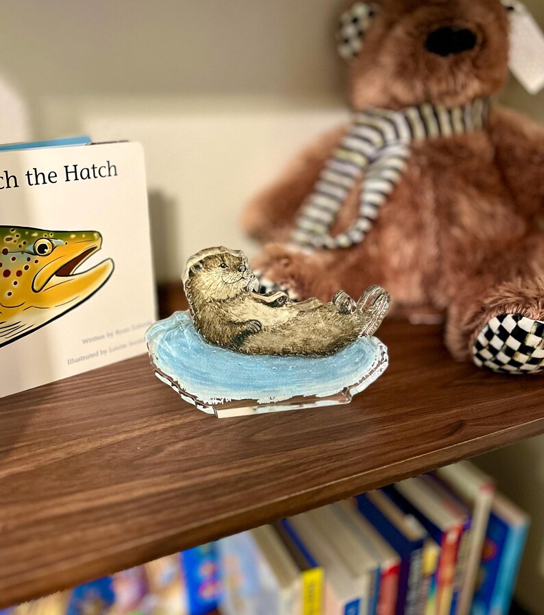 May include: A detailed figurine of a sea otter resting on a light blue base. The otter is brown with a light belly, and the base has a white border. A children's book and a brown teddy bear are in the background.