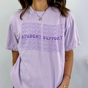 May include: A light purple t-shirt with the words "STUDENT SUPPORT" in a repeating pattern across the front. The text is in a darker shade of purple. The shirt has short sleeves and a crew neck.