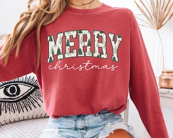 May include: A red long-sleeve sweatshirt with the text "MERRY CHRISTMAS" in a green and white design. The word "MERRY" is in a collegiate-style font, and "christmas" is in a script font. The sweatshirt is paired with denim shorts.