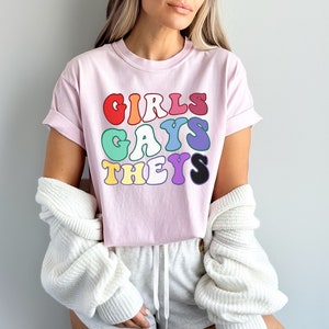 May include: A pink t-shirt with the text "GIRLS GAYS THEYS" in rainbow colors. The text is in a retro font style.