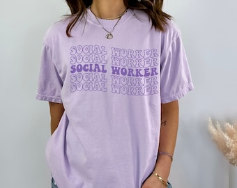 Op de afbeelding: Een lavendelkleurig T-shirt met de tekst "SOCIAL WORKER" die vijf keer wordt herhaald in een paars retro-lettertype.