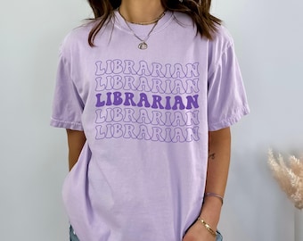 Può includere: Una maglietta color lavanda con la parola "LIBRARIAN" ripetuta cinque volte in un font retrò viola e bianco.