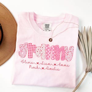 May include: A light pink t-shirt with the word "Grammy" in a patchwork design of pink hearts and polka dots. Below the word are the names Olivia, Liam, Noah, Emma, and Amelia. A gold necklace with a pendant is on the shirt.