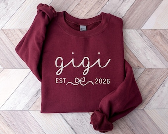 May include: A maroon sweatshirt with the word "gigi" in white script. Below the word "gigi" is the text "EST 2026" with a bow graphic. The sweatshirt is folded on a light brown and white wooden surface.