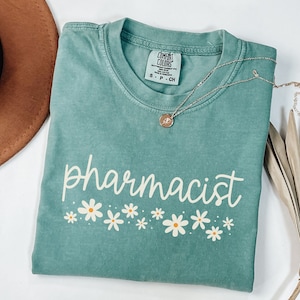 May include: A teal blue t-shirt with the text "pharmacist" in white script with a row of white daisies underneath. The shirt is made of a soft, comfortable material.