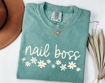 May include: A teal blue t-shirt with the text "nail boss" in white script with a row of white daisies with yellow centers below the text.