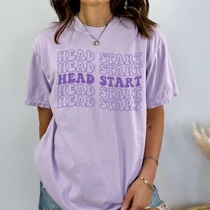 May include: A lavender t-shirt with the text "HEAD START" repeated five times in a purple and white retro font.
