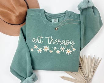 Op de afbeelding: Een tealkleurige sweatshirt met de woorden "art therapy" in witte letters, vergezeld van een rij witte madeliefjes met gele centra. De sweatshirt heeft opgerolde mouwen en een ronde hals. Een gouden ketting met een kleine hanger is zichtbaar.