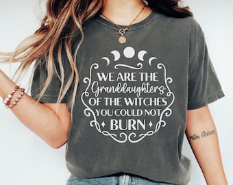 Pode incluir: Uma camiseta cinza com um gráfico branco que diz "We are the Granddaughters of the Witches you could not BURN". O gráfico é cercado por uma borda decorativa e inclui três luas crescentes.