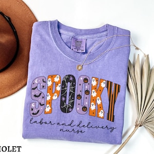 May include: A violet short-sleeved t-shirt featuring the word SPOOKY in large, patterned letters with ghosts, bats, pumpkins, and spiderwebs. Below the word, the text reads labor and delivery nurse in a black cursive font. The shirt has a crew neck.