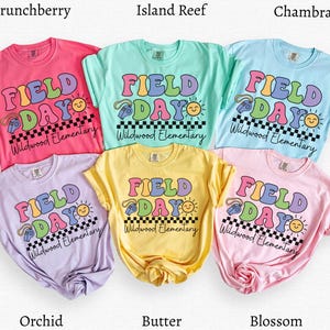 May include: Six short-sleeved t-shirts in various colors, including pink, blue, and yellow. Each shirt features the words "FIELD DAY" in a colorful, retro font, along with a sun and a whistle graphic. The text "Wildwood Elementary" is also printed on each shirt.