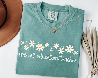 May include: A teal Comfort Colors t-shirt with the words "special education teacher" in white script, adorned with a row of white and orange daisies. A silver necklace with a pendant rests on the shirt.