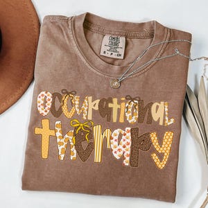 May include: A brown Comfort Colors t-shirt with the words "Occupational Therapy" in a decorative font. The letters are a mix of patterns, including pumpkins, leaves, and stripes. A silver necklace with a pendant is draped across the shirt.