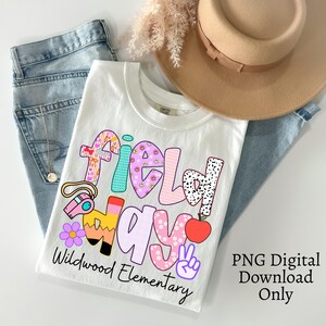 Pode incluir: Camiseta branca com um gráfico colorido "Field Day", apresentando um apito, um lápis, uma maçã e um sinal de paz. O texto "Wildwood Elementary" está abaixo do gráfico. A camisa é combinada com jeans azul e um chapéu bege.
