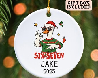 May include: White ceramic Christmas ornament featuring a cartoon goose wearing sunglasses, a Santa hat, and a Christmas sweater. The ornament has the text "SIX-SEVEN JAKE 2025" in red and black. A gold string is attached for hanging.