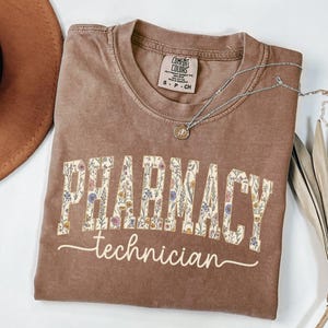 May include: A brown t-shirt with the words "PHARMACY technician" in a floral design. The shirt has a crew neck and a small tag on the upper back. A silver necklace with a pendant is draped across the shirt.