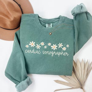 May include: A green sweatshirt with a row of white daisies and the text "cardiac sonographer" in white.