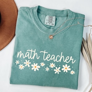 May include: A teal blue t-shirt with the text "math teacher" in white script with a row of white daisies with yellow centers below the text.