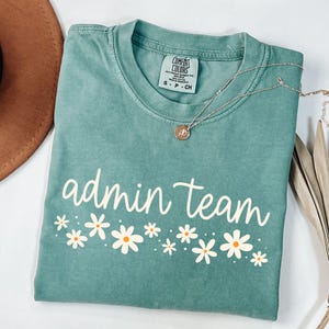 May include: A teal blue t-shirt with the text "admin team" in white with a row of white daisies with yellow centers below the text.